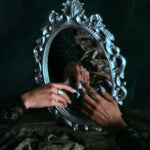 Models hands with wearing 18k hand-engraved diamond moon ring along with other jewelry in front of a baroque mirror and dark backdrop.