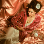 Model with gold heart necklace and gold ring reposing in front of a pink silk backdrop.