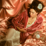 Model with gold heart necklace and gold ring reposing in front of a pink silk backdrop.