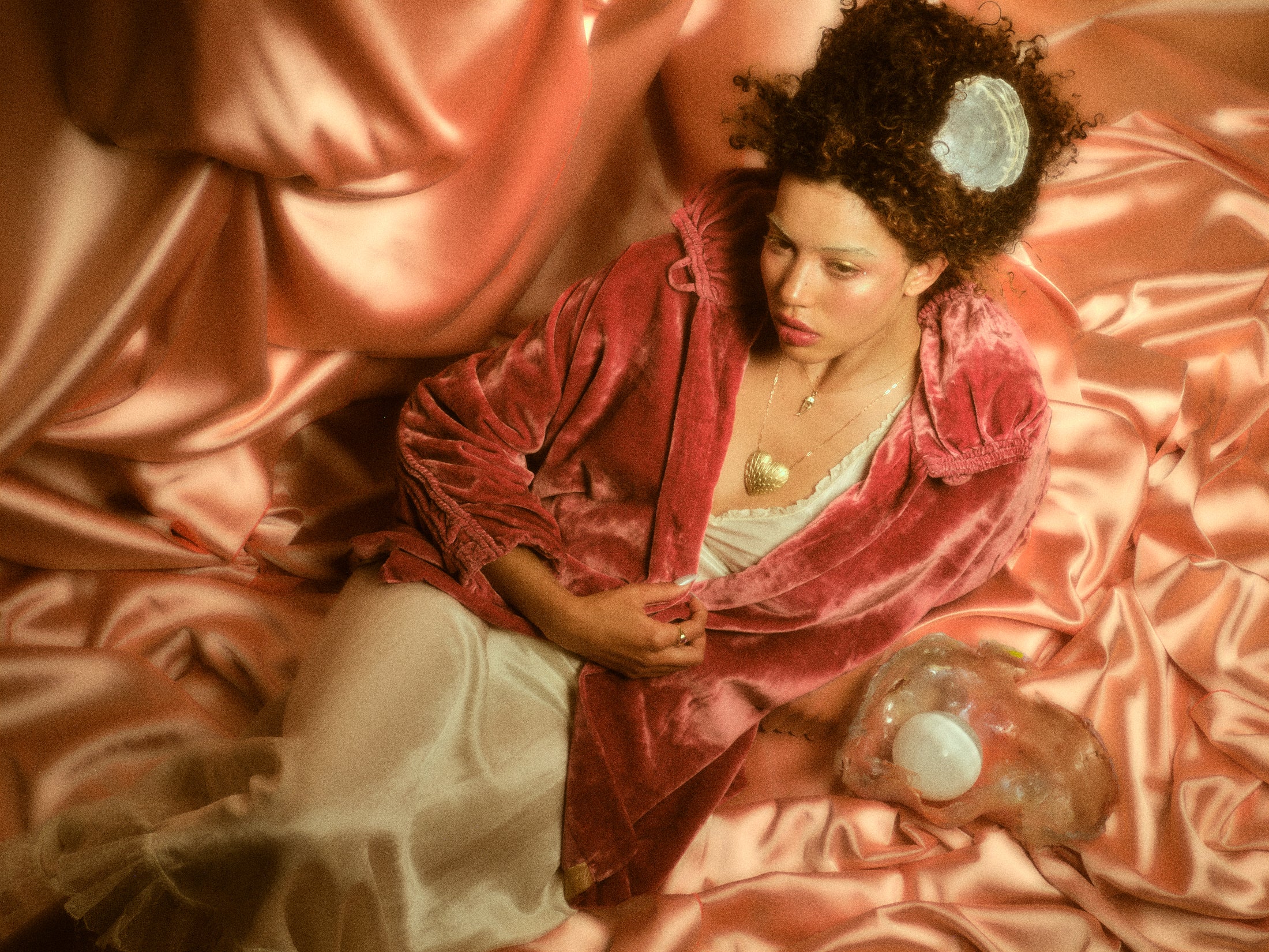 Model with gold heart necklace and gold ring reposing in front of a pink silk backdrop.