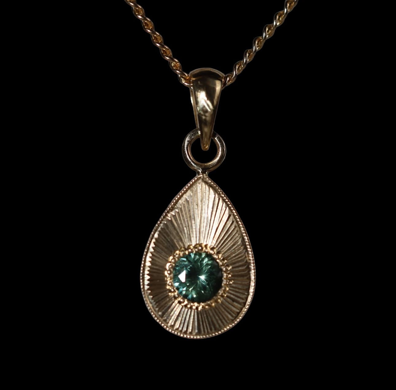 Gold teardrop pendant with a large blue green sapphire on a black background.