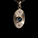 Front view of 14k yellow gold portal charm.  Tall oval shaped with central set blue sapphire, white diamonds set top and bottom and engraved lines radiating to the left and right.  Attached to separately available yellow gold chain.