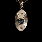 Front view of 14k yellow gold portal charm.  Tall oval shaped with central set blue sapphire, white diamonds set top and bottom and engraved lines radiating to the left and right.  Attached to separately available yellow gold chain.