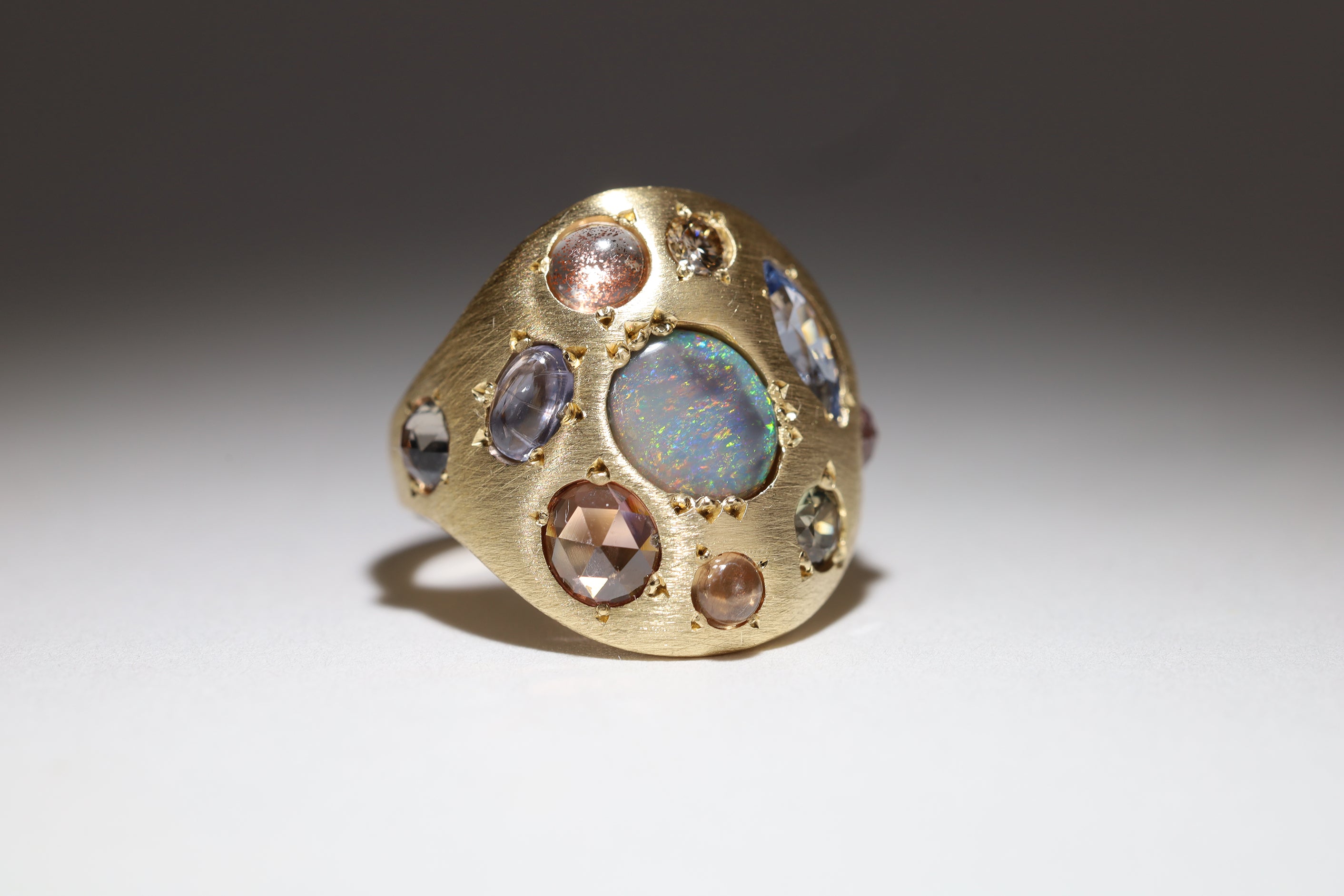 Alternate three quarter view of 18k yellow gold gem palette ring with ten unique gems.  Showing a central semi black Australian opal surrounded by a dusty blue marquise sapphire, pale green sapphire, rose colored sapphire cabochon, rose-cut burnt peach zircon, lavender sapphire cabochon, a sunstone, champagne diamond, pale blue rose cut sapphire, and pink rose cut sapphire.