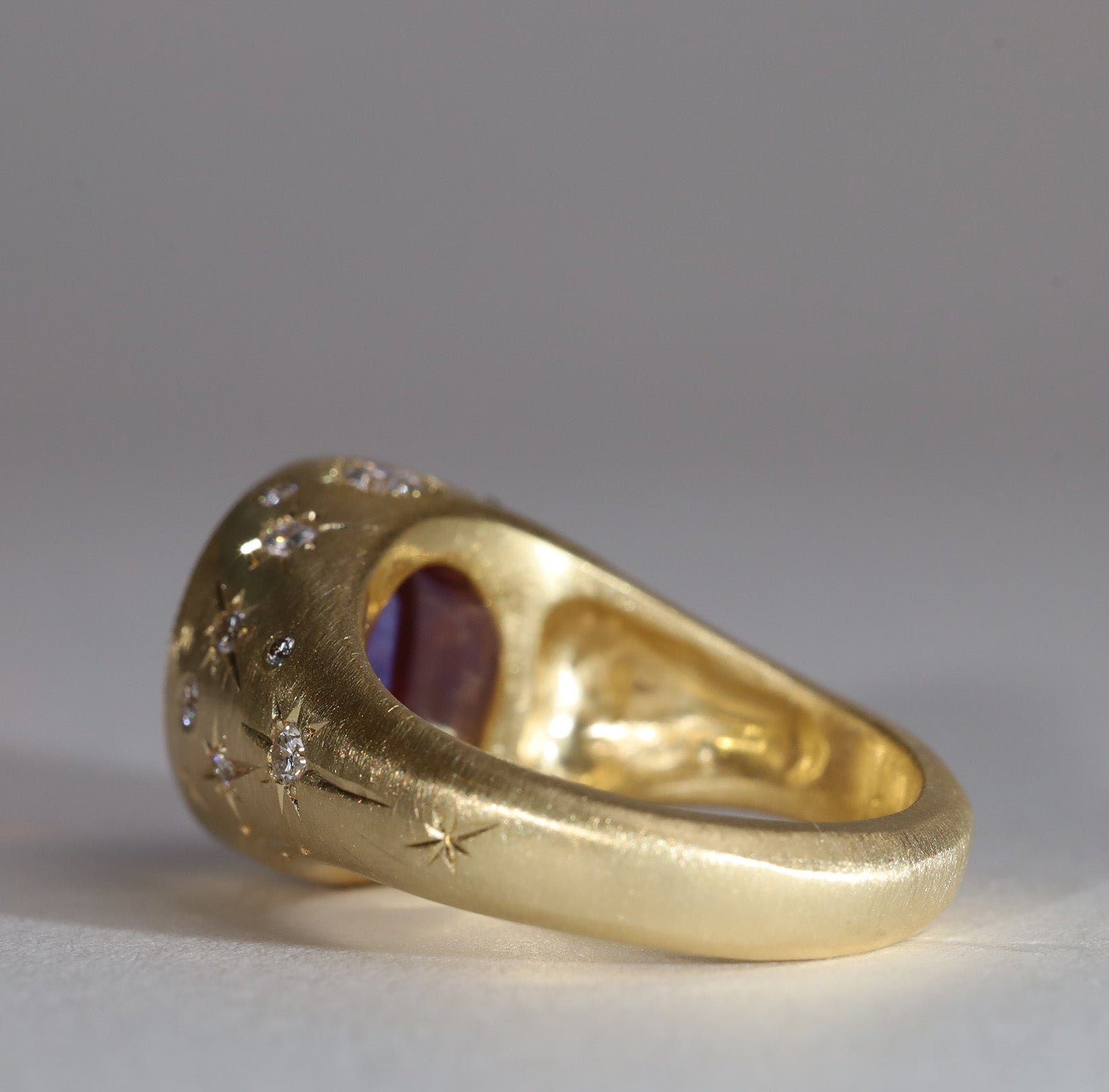 Gold signet style ring with large central purple sapphire, engraved with stars and comets, some set with diamonds. Rear view facing left.