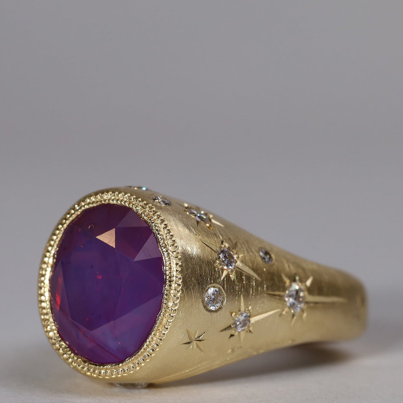 Gold signet style ring with large central purple sapphire, engraved with stars and comets, some set with diamonds. Side view facing left.
