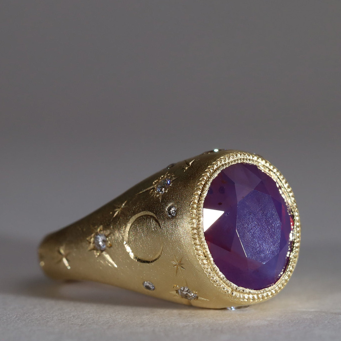 Gold signet style ring with large central purple sapphire, engraved with stars and comets, some set with diamonds.  Side view facing right.