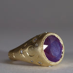 Gold signet style ring with large central purple sapphire, engraved with stars and comets, some set with diamonds.  Side view facing right.