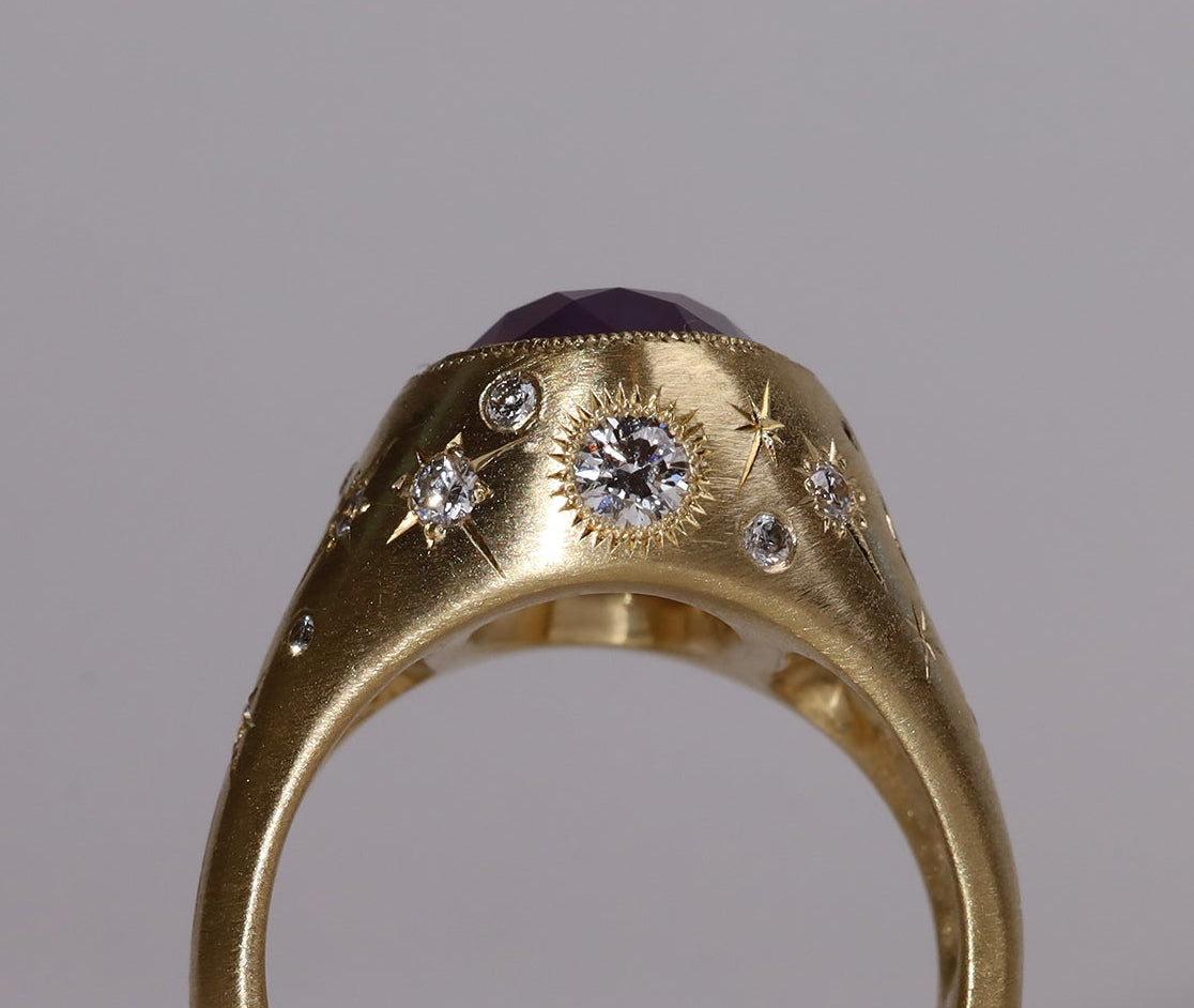Gold signet style ring with large central purple sapphire, engraved with stars and comets, some set with diamonds.  Standing view wide side 1