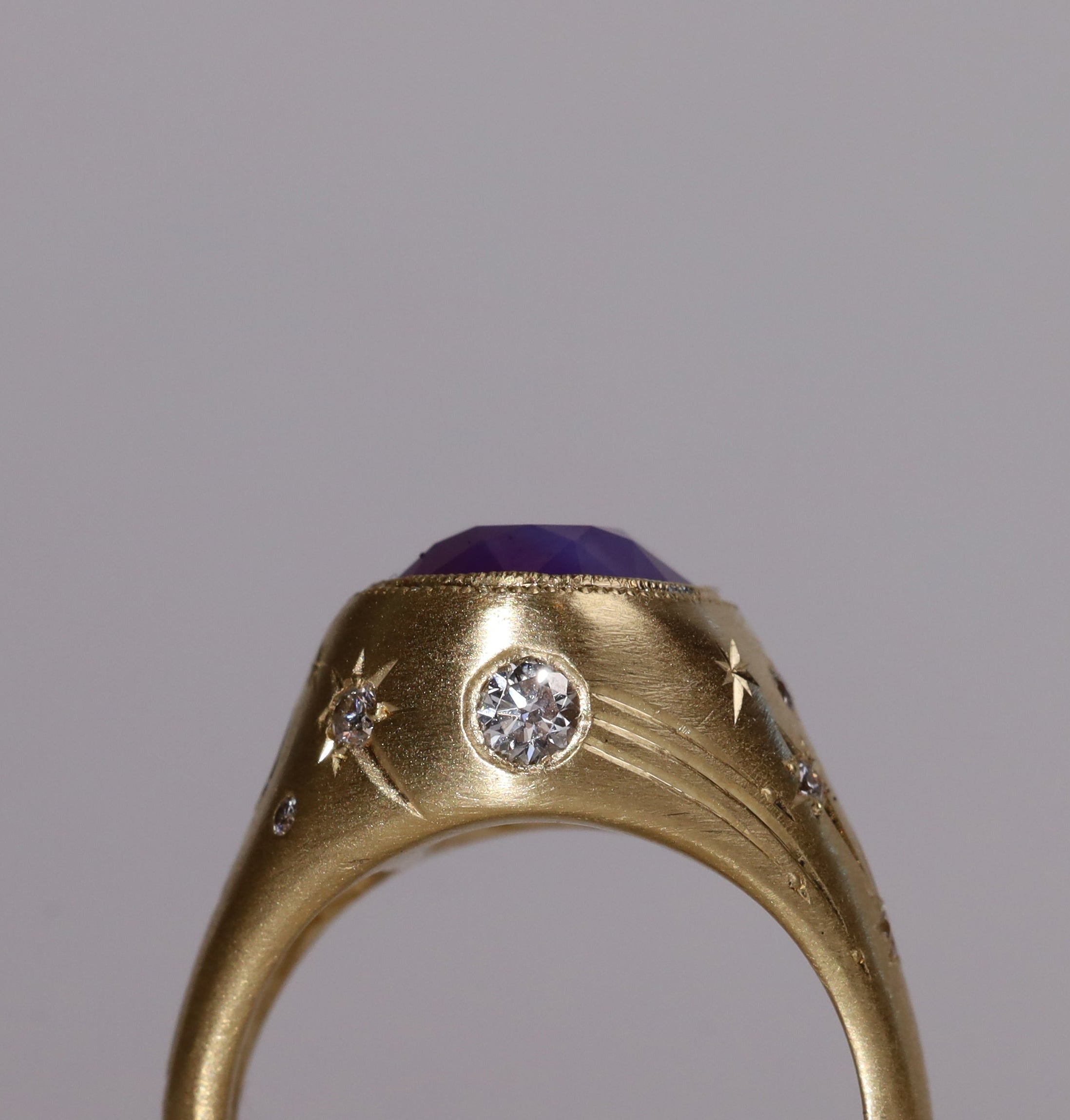 Gold signet style ring with large central purple sapphire, engraved with stars and comets, some set with diamonds.  Standing view alternate wide side