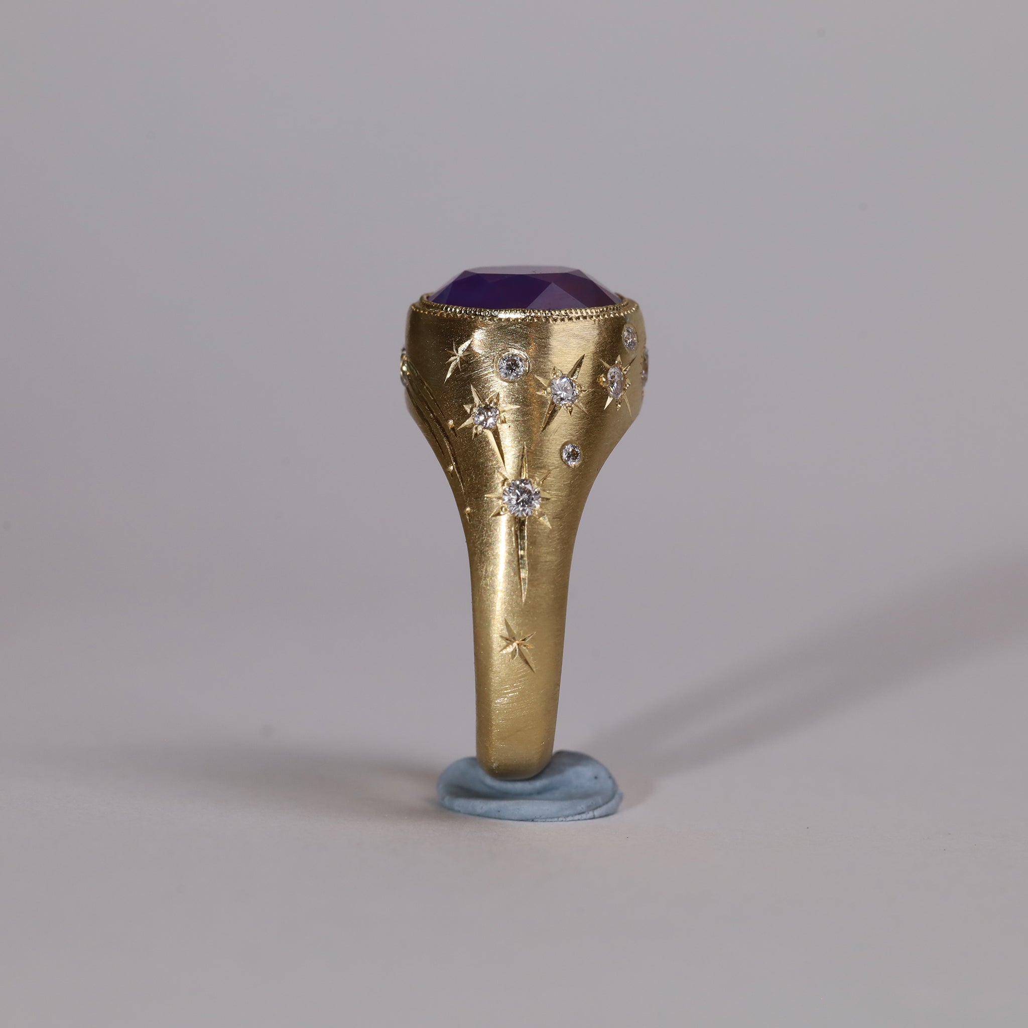 Gold signet style ring with large central purple sapphire, engraved with stars and comets, some set with diamonds.  Standing view narrow side 1