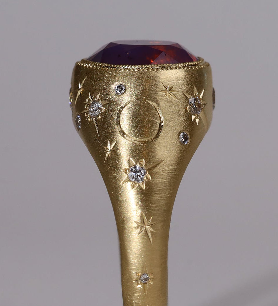Gold signet style ring with large central purple sapphire, engraved with stars and comets, some set with diamonds.  Standing view alternate narrow side.