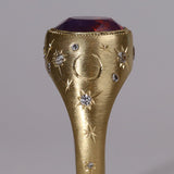 Gold signet style ring with large central purple sapphire, engraved with stars and comets, some set with diamonds.  Standing view alternate narrow side.