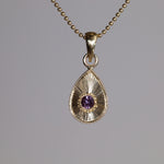 Gold teardrop pendant with a purple gemstone on a gray background