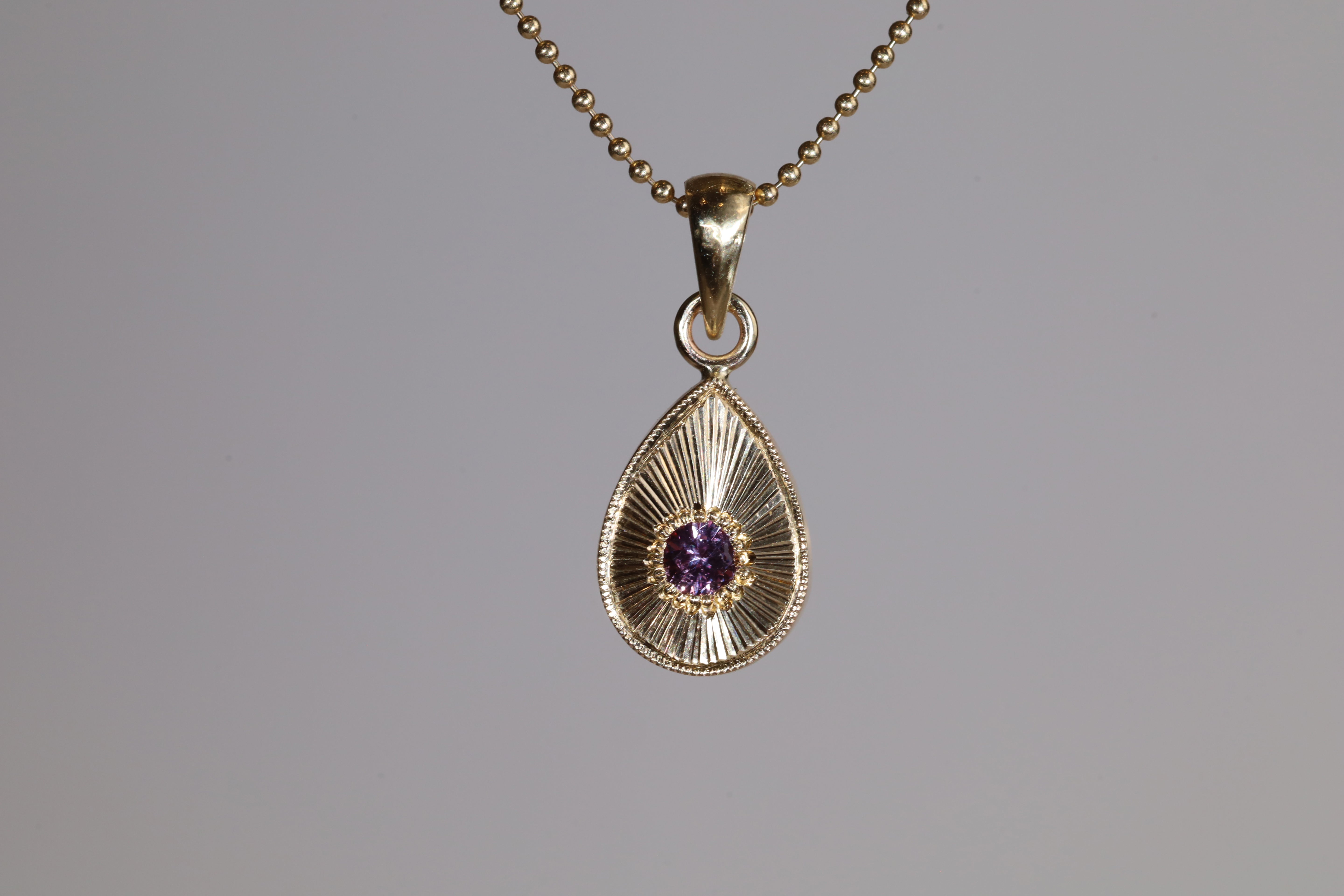 Gold teardrop pendant with a purple gemstone on a gray background