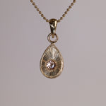 Gold teardrop pendantt with a pink and peach stone on a grey background.
