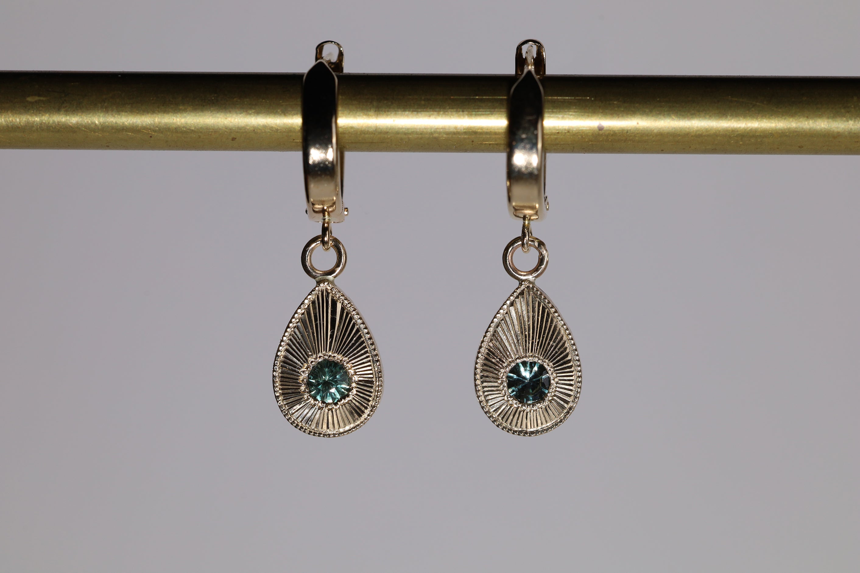 Front view of 14k yellow gold plume earrings, which feature a tear drop shape with engraved rays emanating from central aqua sapphire.