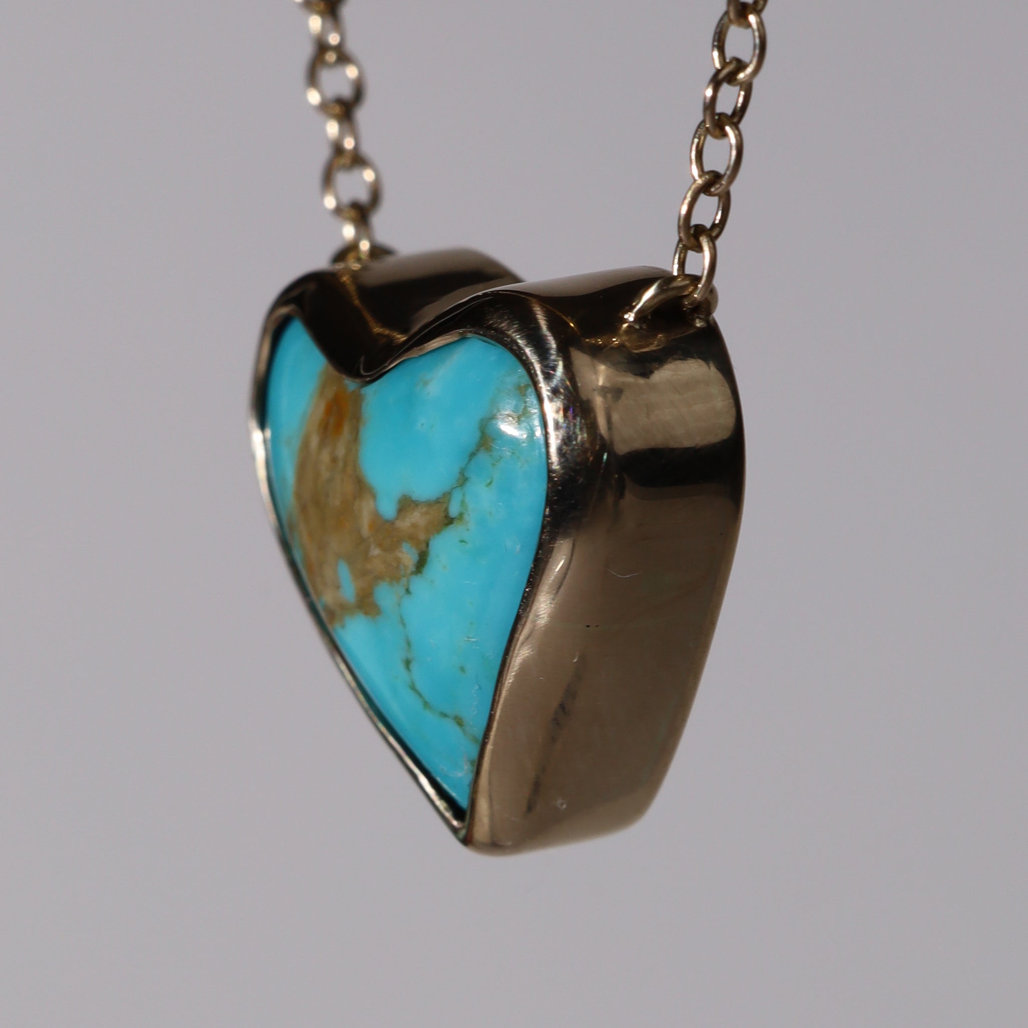 Three quarter side view of a 14k Pendant with a heart shaped Kingman Turquoise stone.  Hanging in front of a flat grey background with its chain extending out of frame.