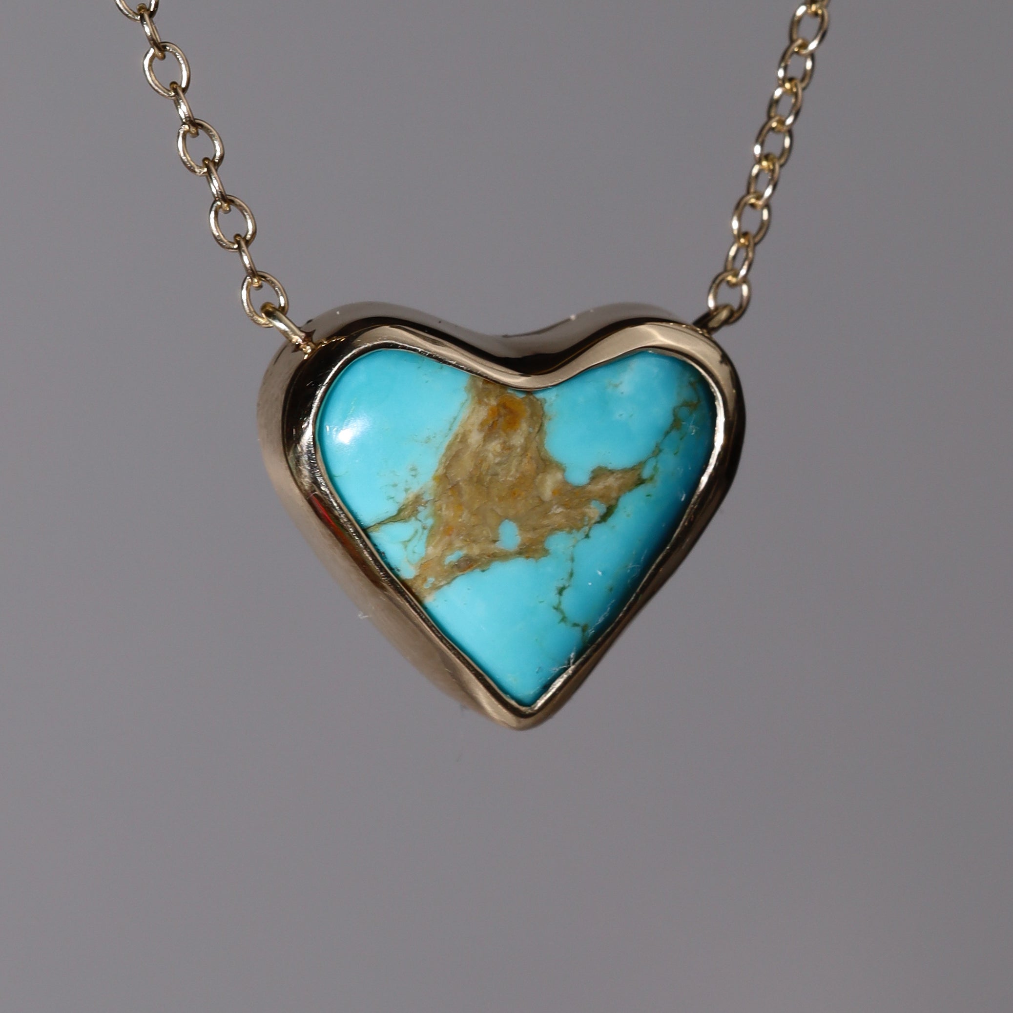 14k Pendant with a heart shaped Kingman Turquoise stone.  Hanging in front of a flat grey background with its chain extending out of frame.